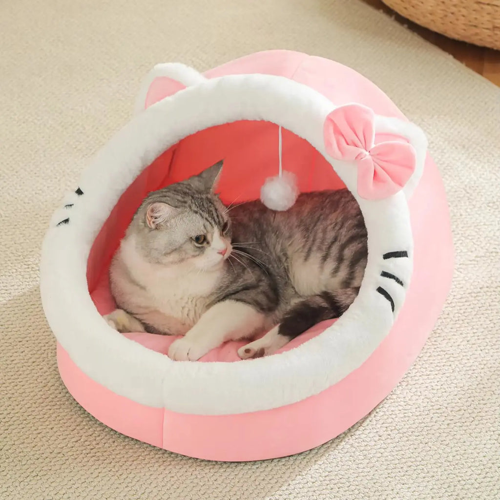 Pink and white cat-face shaped Cosy Cave Cat Bed with bow accent, two grey tabby cats resting inside on a beige rug