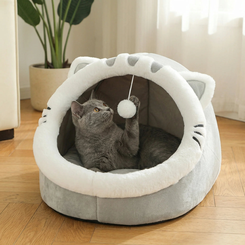 Grey cat-face shaped Cosy Cave Cat Bed with a grey cat batting at the hanging pom-pom toy inside, bright living room with a potted plant