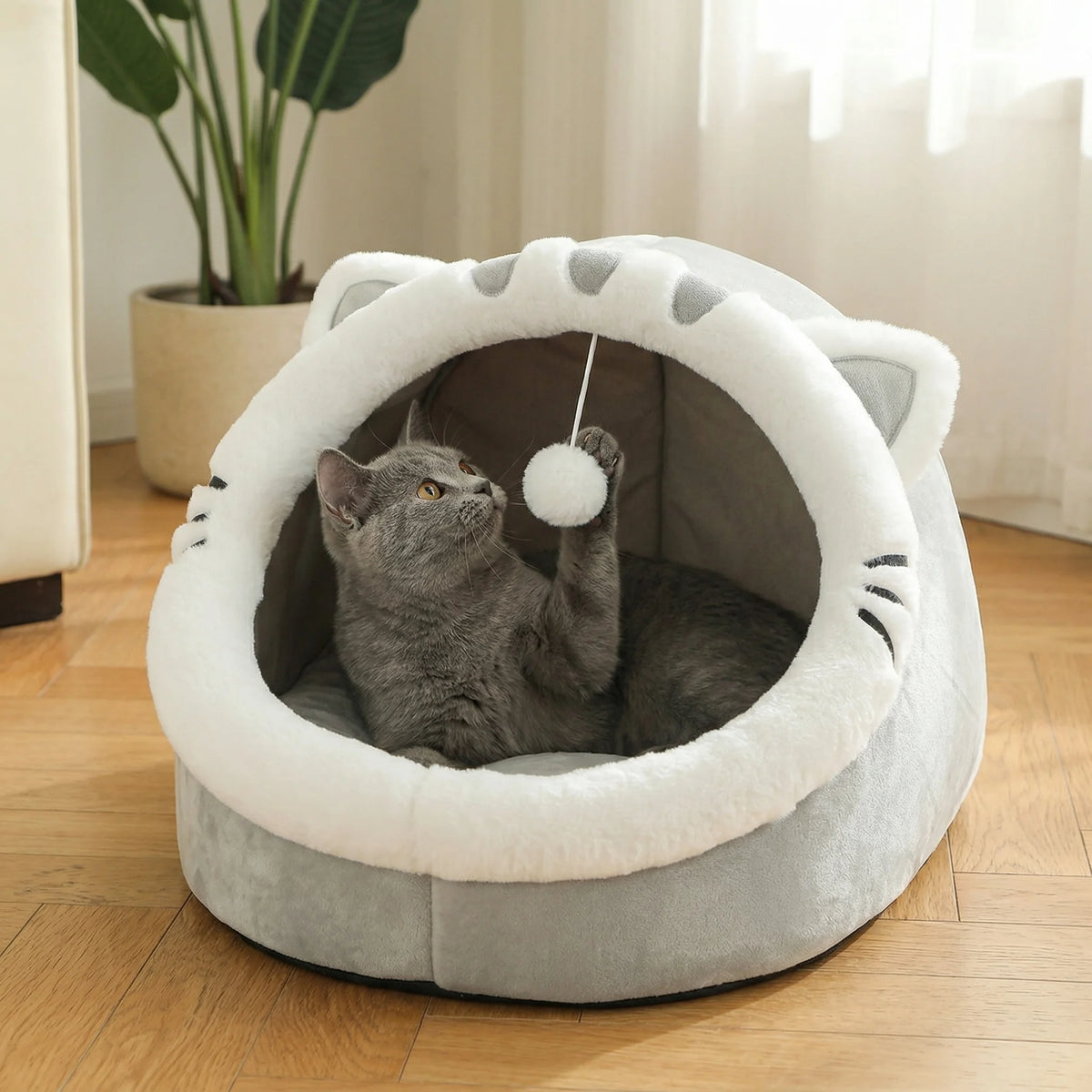 Grey cat-face shaped Cosy Cave Cat Bed with a grey cat batting at the hanging pom-pom toy inside, bright living room with a potted plant