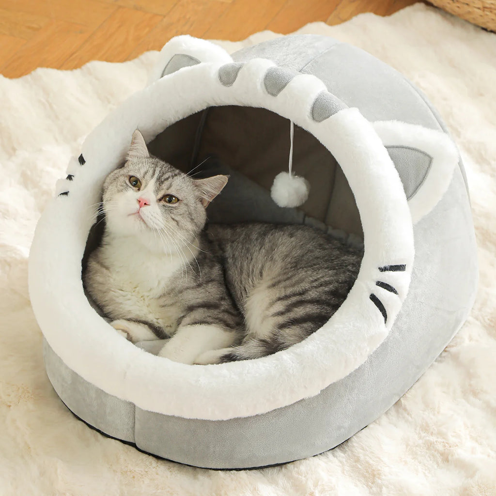 Grey cat-face shaped Cosy Cave Cat Bed with a grey and white cat looking up while lying inside on a white fluffy rug