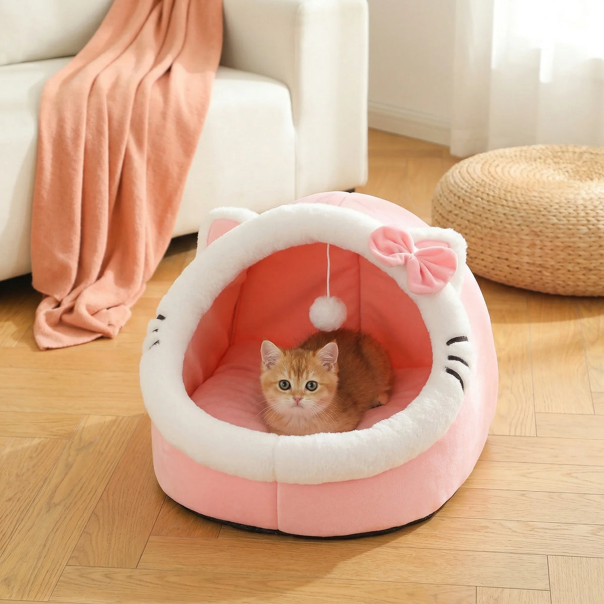 Pink cat-face shaped Cosy Cave Cat Bed with bow accent, ginger kitten sitting inside near a white armchair with a blush throw blanket